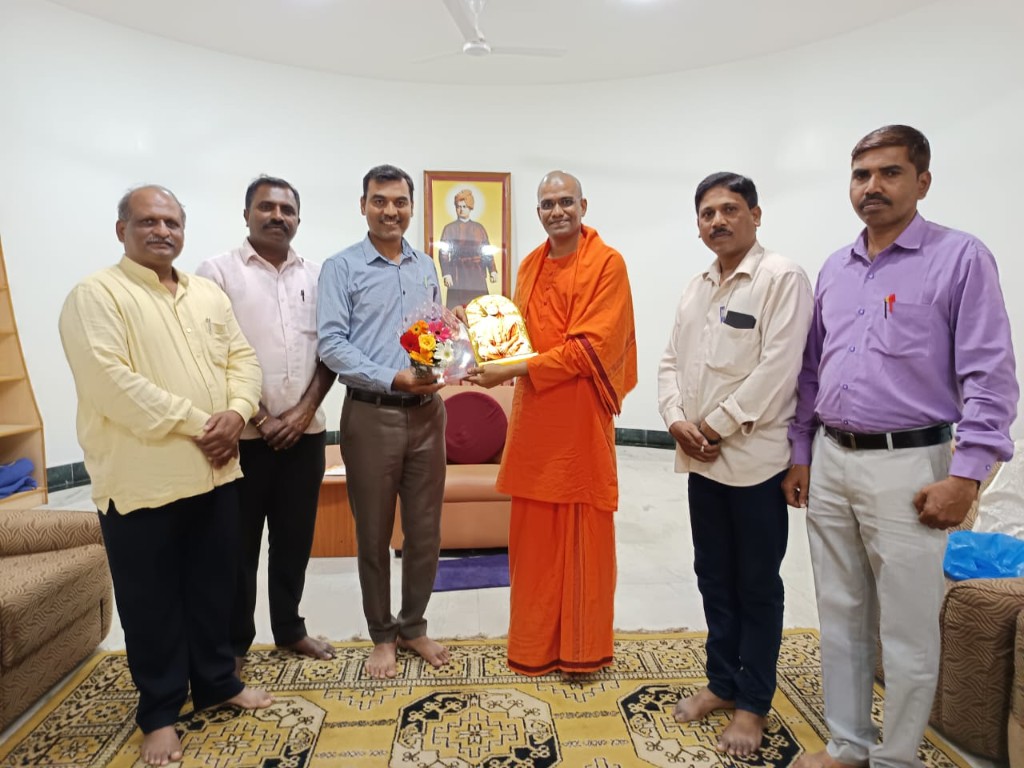 Rajashekhar Ragati presenting flowers to a monk in orange robes, with colleagues, indoors